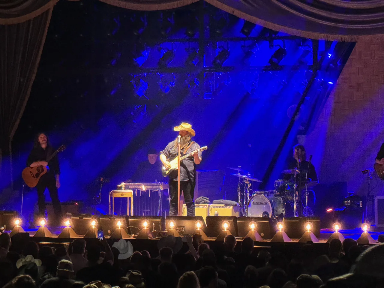 Chris Stapleton singing and playing guitar under a warm spotlight against a deep blue stage, with his band beside him.