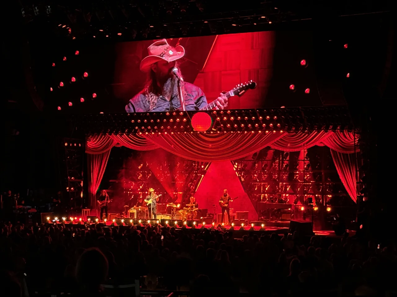 Chris Stapleton and band framed by red curtains and a large video close-up above the stage.