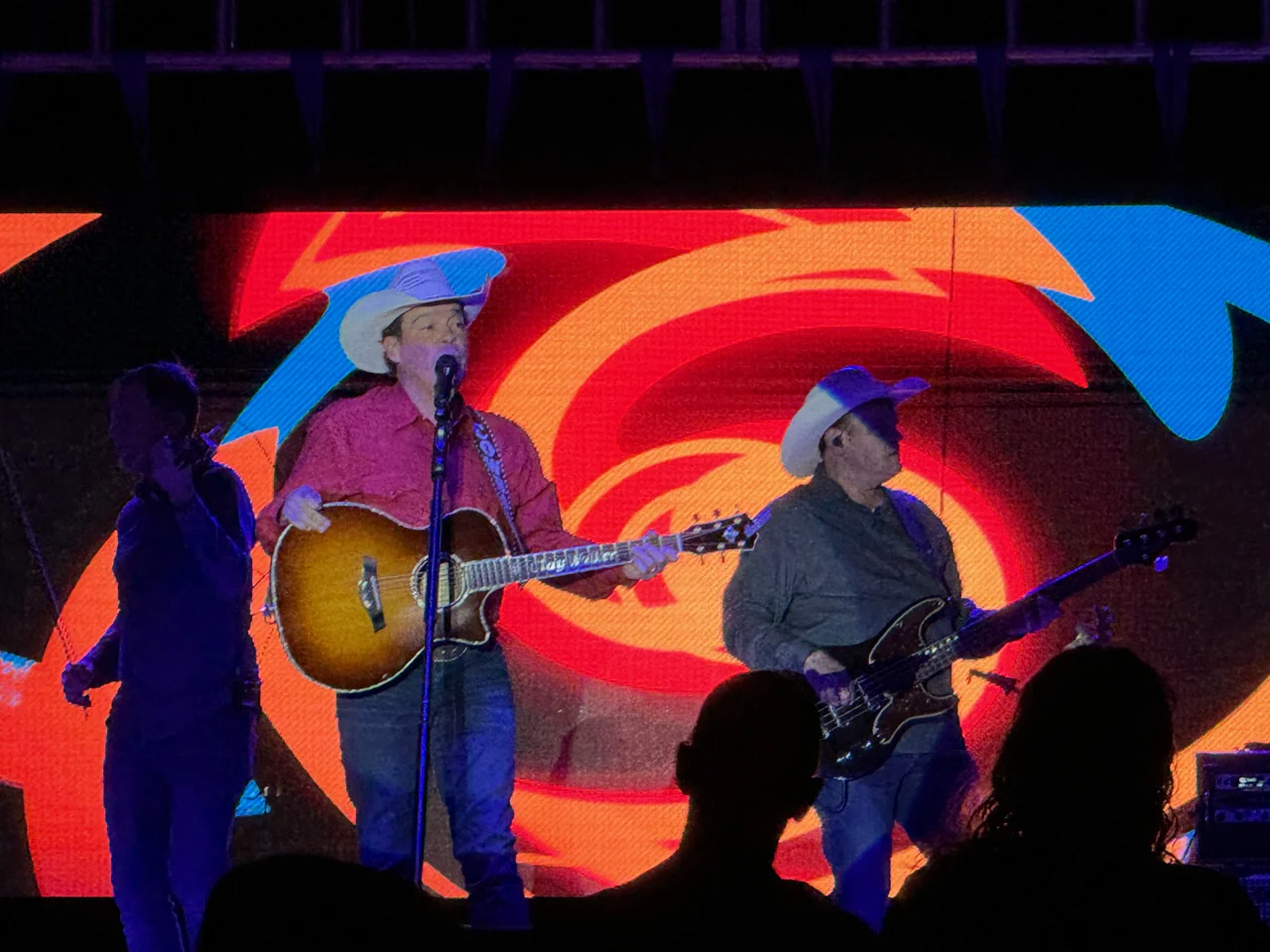 Clay Walker in a red shirt and white cowboy hat, playing guitar before a red, orange, and blue spiral backdrop, with audience silhouettes in front.