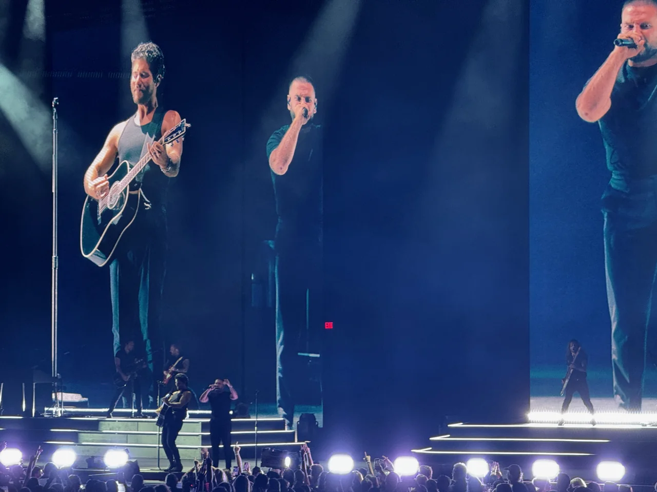 Dan + Shay performing between tall video screens showing enlarged views of both singers under blue light.
