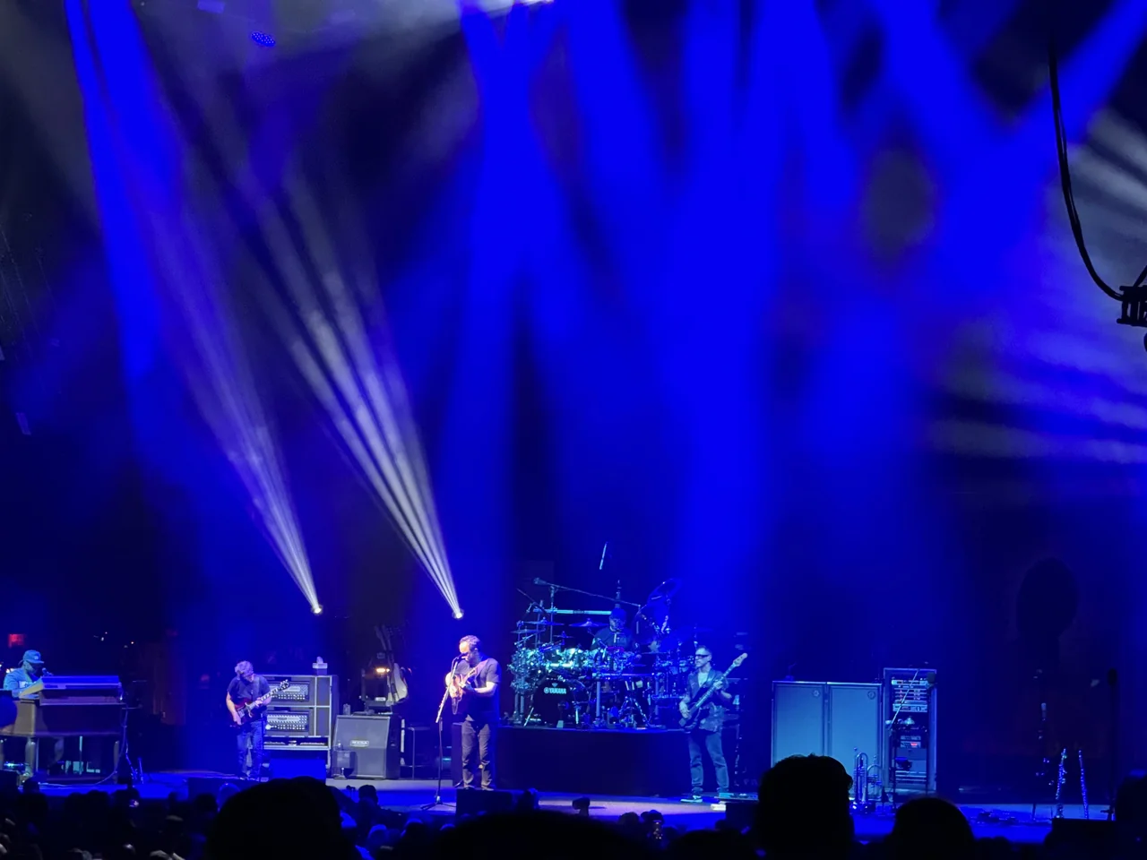 Dave Matthews Band spread across a blue-lit stage beneath fans of white light, with audience silhouettes below.
