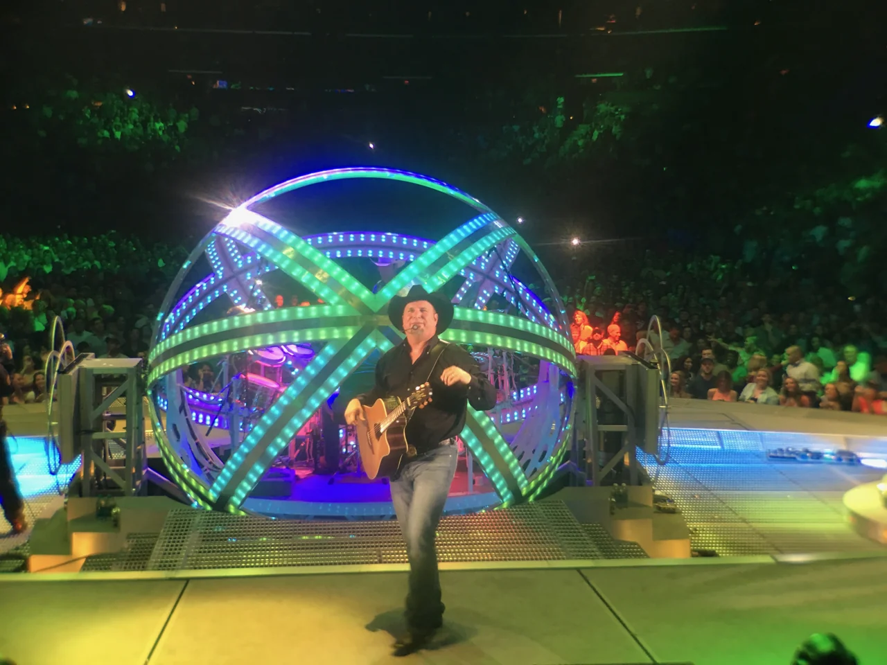Garth Brooks smiling with an acoustic guitar in front of a spherical drum enclosure illuminated in green and blue, with audience seating behind the stage.