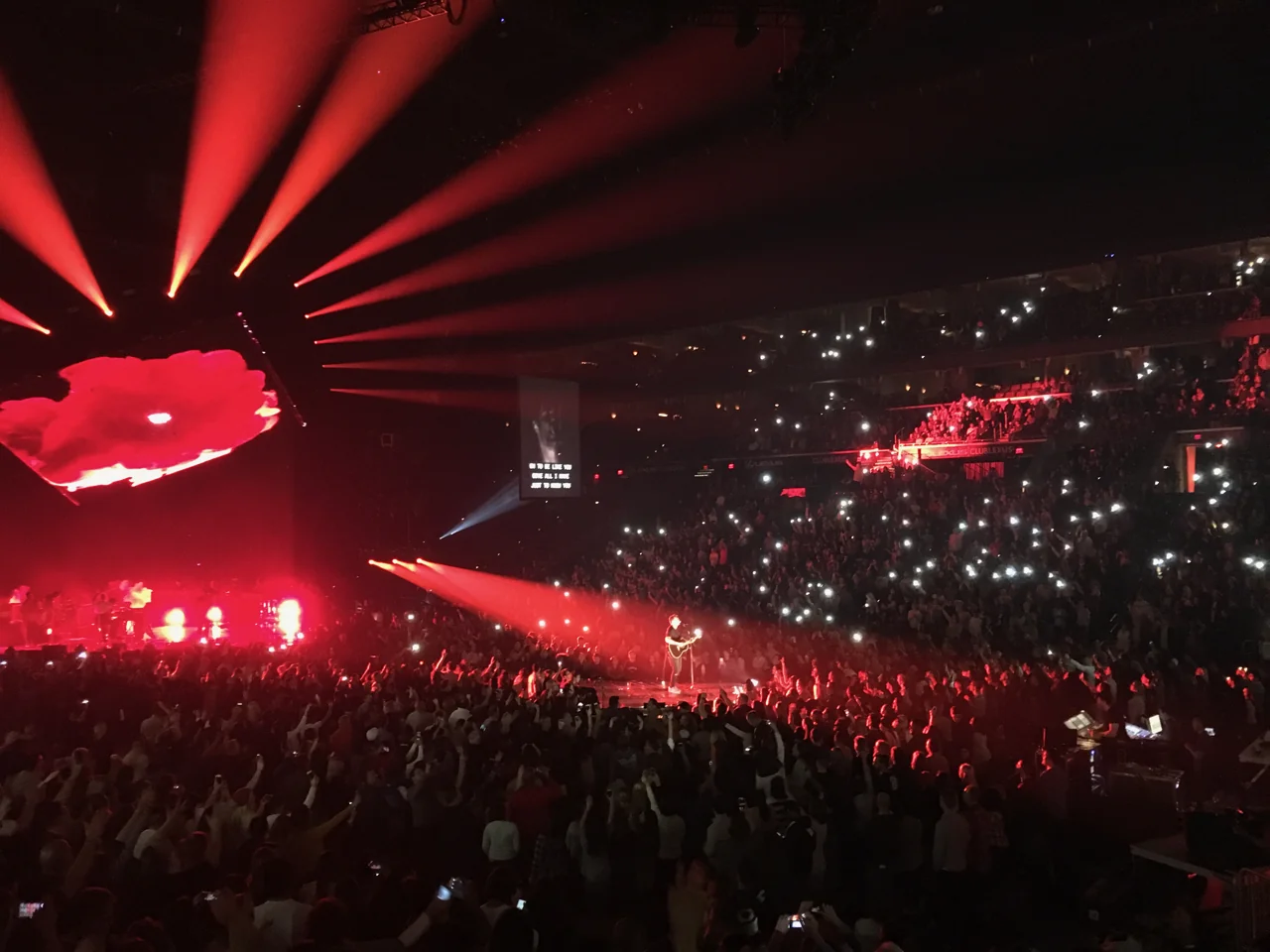 Hillsong UNITED performing on the main stage and a platform surrounded by the audience, beneath red beams and a tilted screen, with phone lights throughout the arena.
