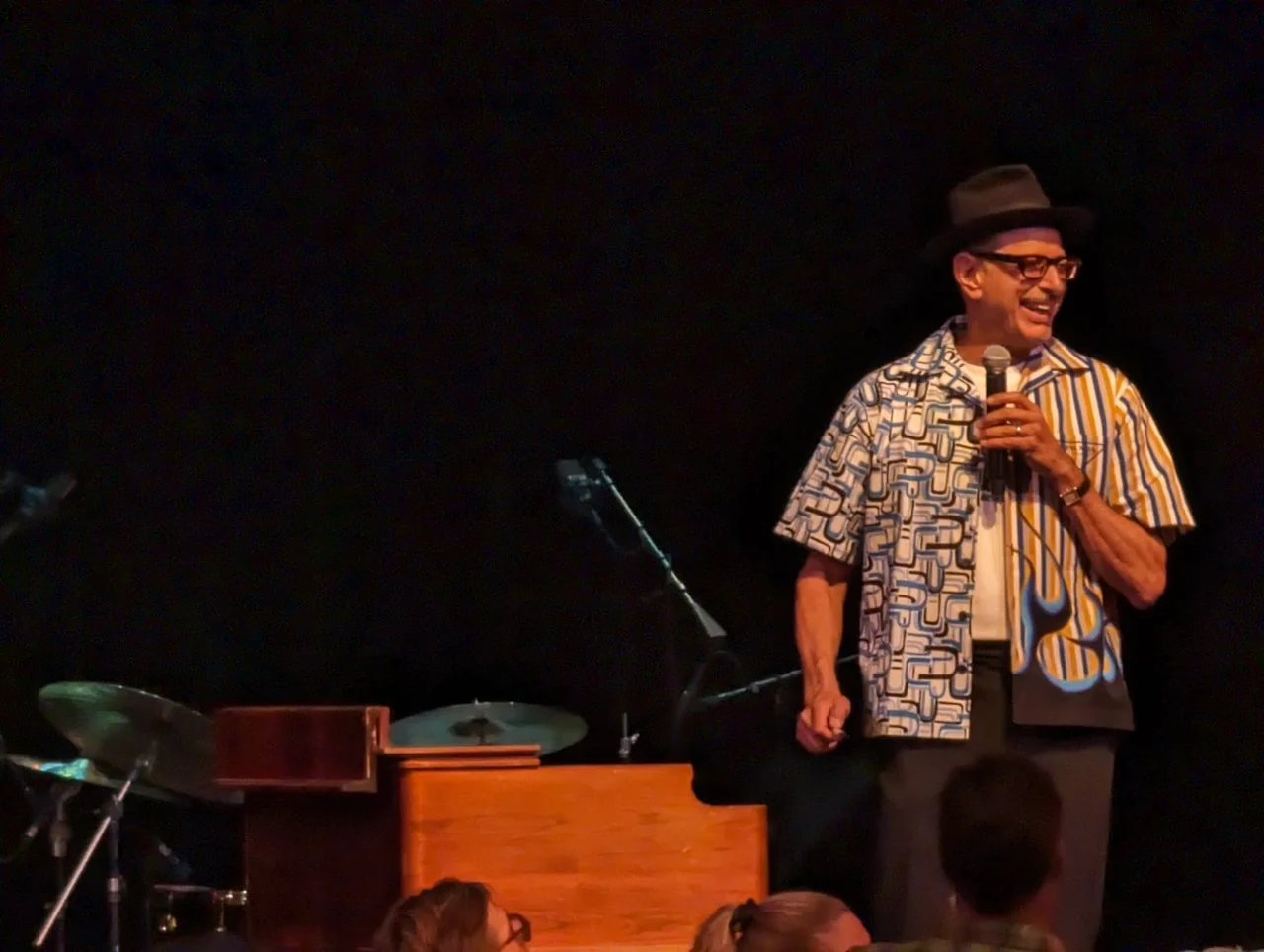 Jeff Goldblum smiling with a handheld microphone beside the piano.