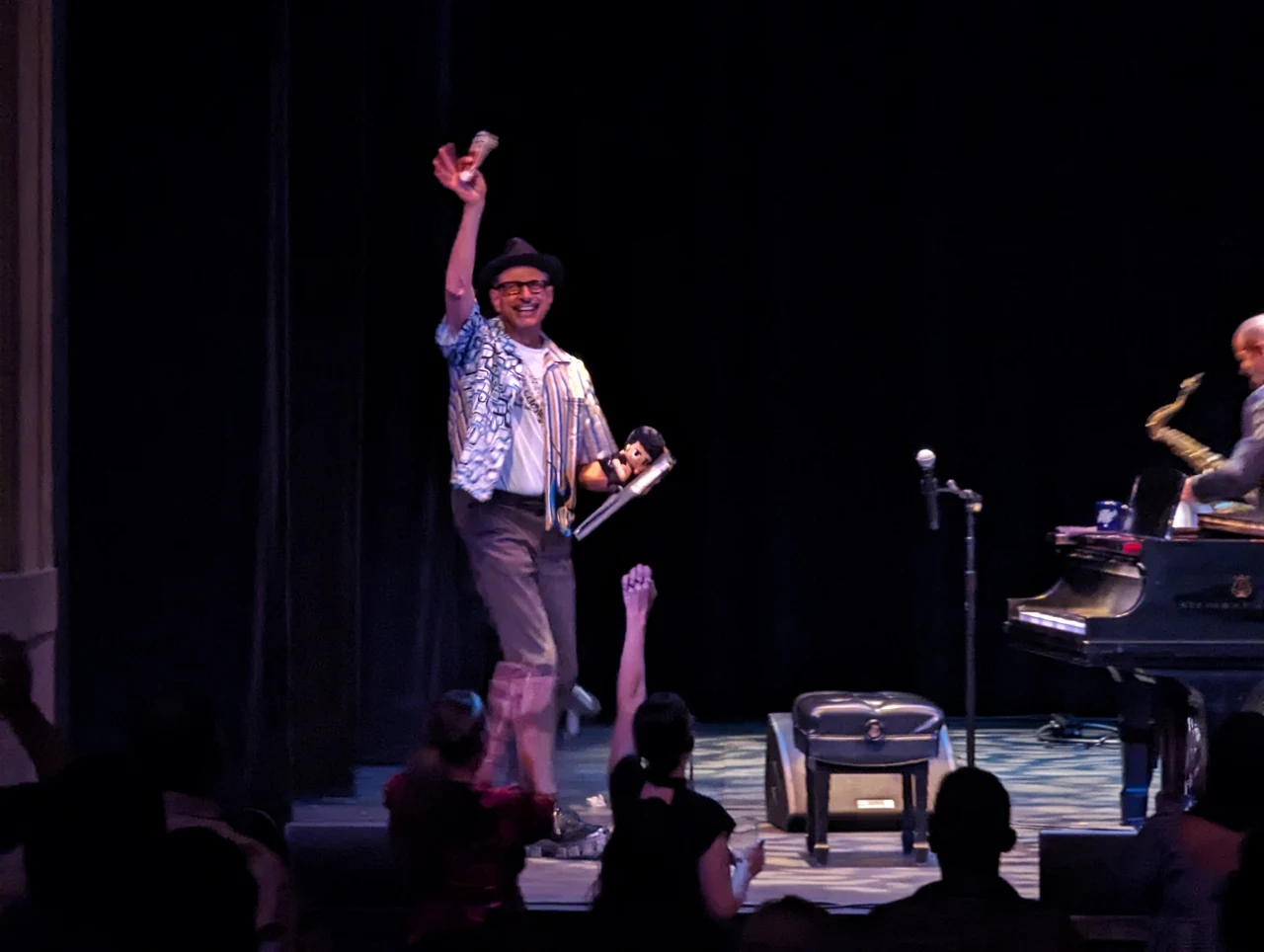Jeff Goldblum smiling with one hand raised toward the audience, beside a Steinway piano and a saxophonist at Balboa Theatre.