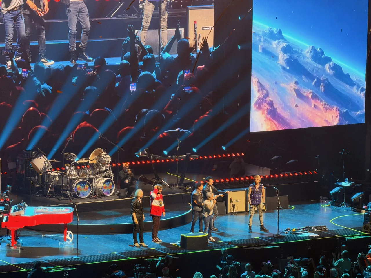 Journey members gathered at the front of a blue-lit stage, one wearing a red Florida Panthers jersey, with a red piano to the left and a large video screen above.