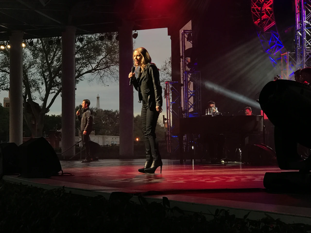 Kerry Butler singing in the foreground and Kevin Massey farther left on Epcot’s open-air stage, with musicians at the piano behind them.