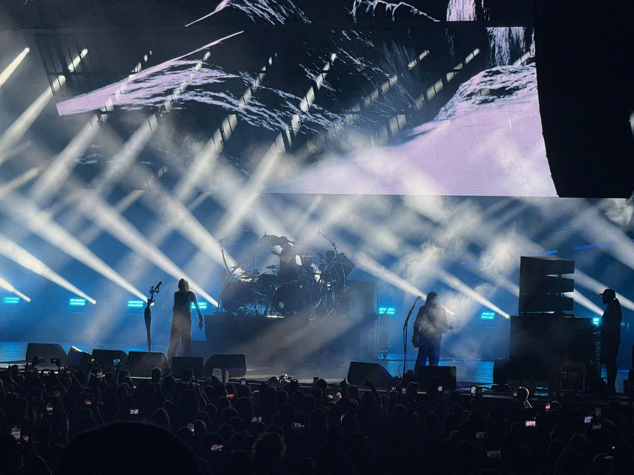 Korn silhouetted beneath intersecting white stage beams and monochrome video screens.