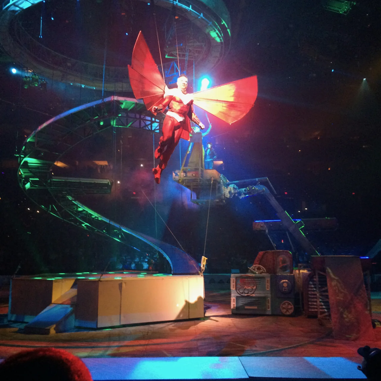 An aerial performer portraying Falcon, with large red wings, suspended above the Marvel Universe LIVE! set amid curved ramps and blue-green lighting.