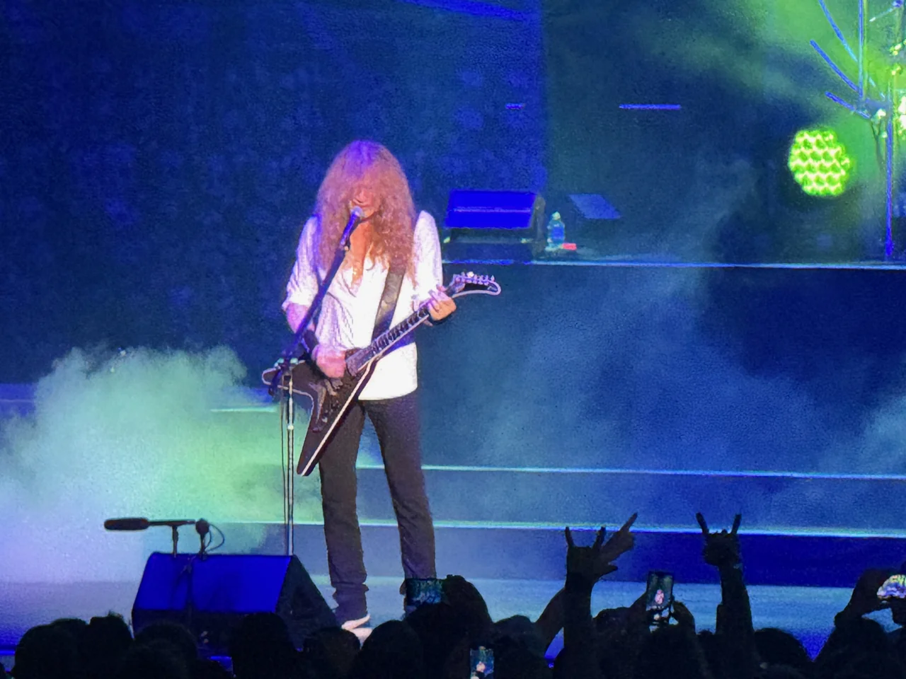 Dave Mustaine playing a black V-shaped guitar under blue and green stage lights, with raised audience hands making horn gestures in the foreground.