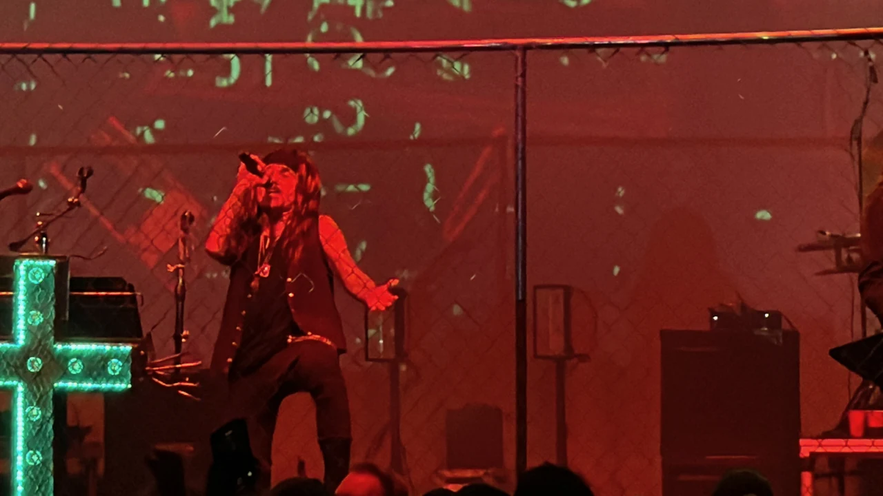 Al Jourgensen singing behind a chain-link fence under red light, with one knee raised, his free hand extended, and an illuminated cross to the left.