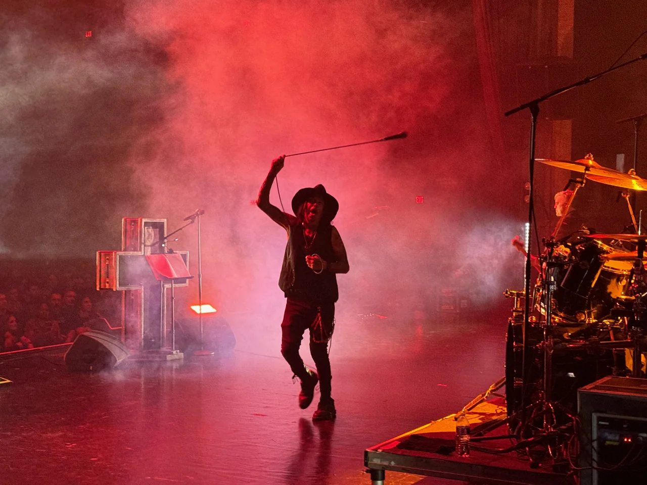 Al Jourgensen holding a microphone stand overhead in red stage haze.