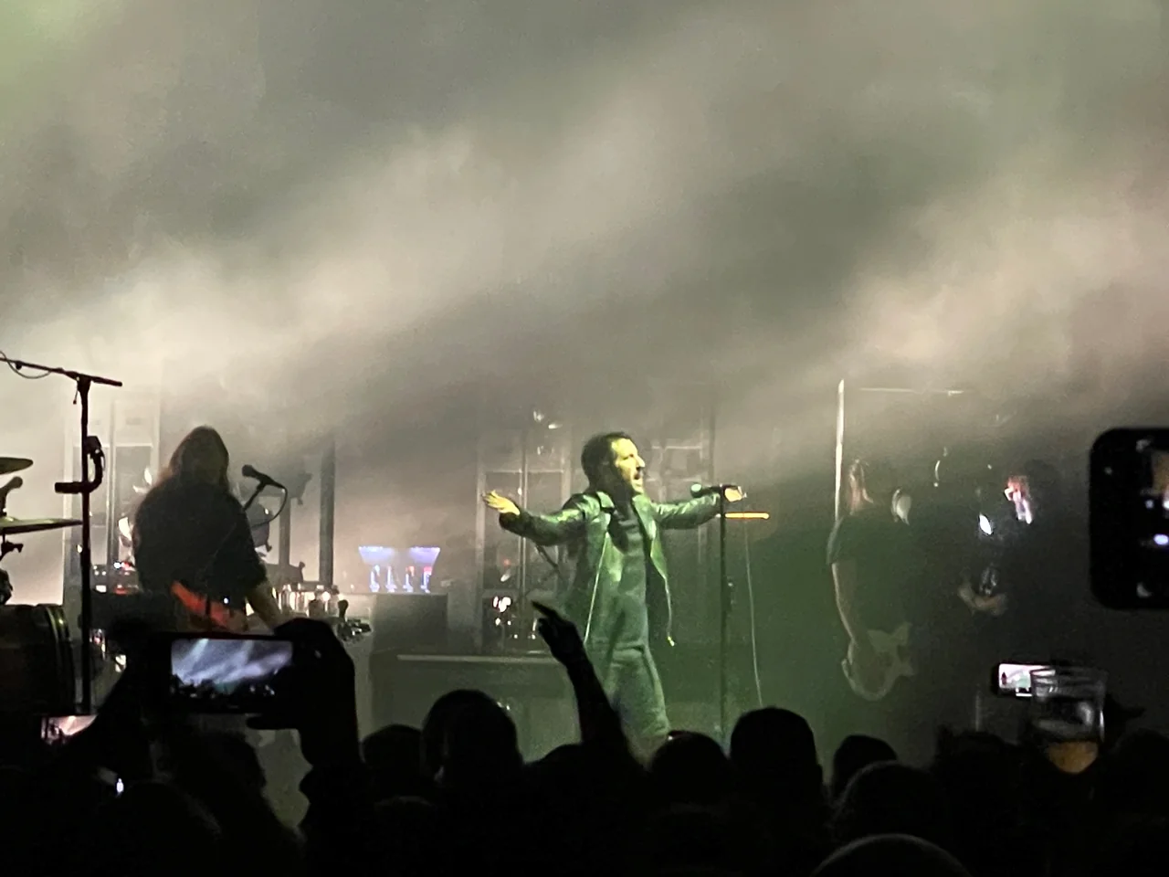 Trent Reznor standing with both arms extended at the microphone, framed by his band, pale stage haze, and audience phones in silhouette.