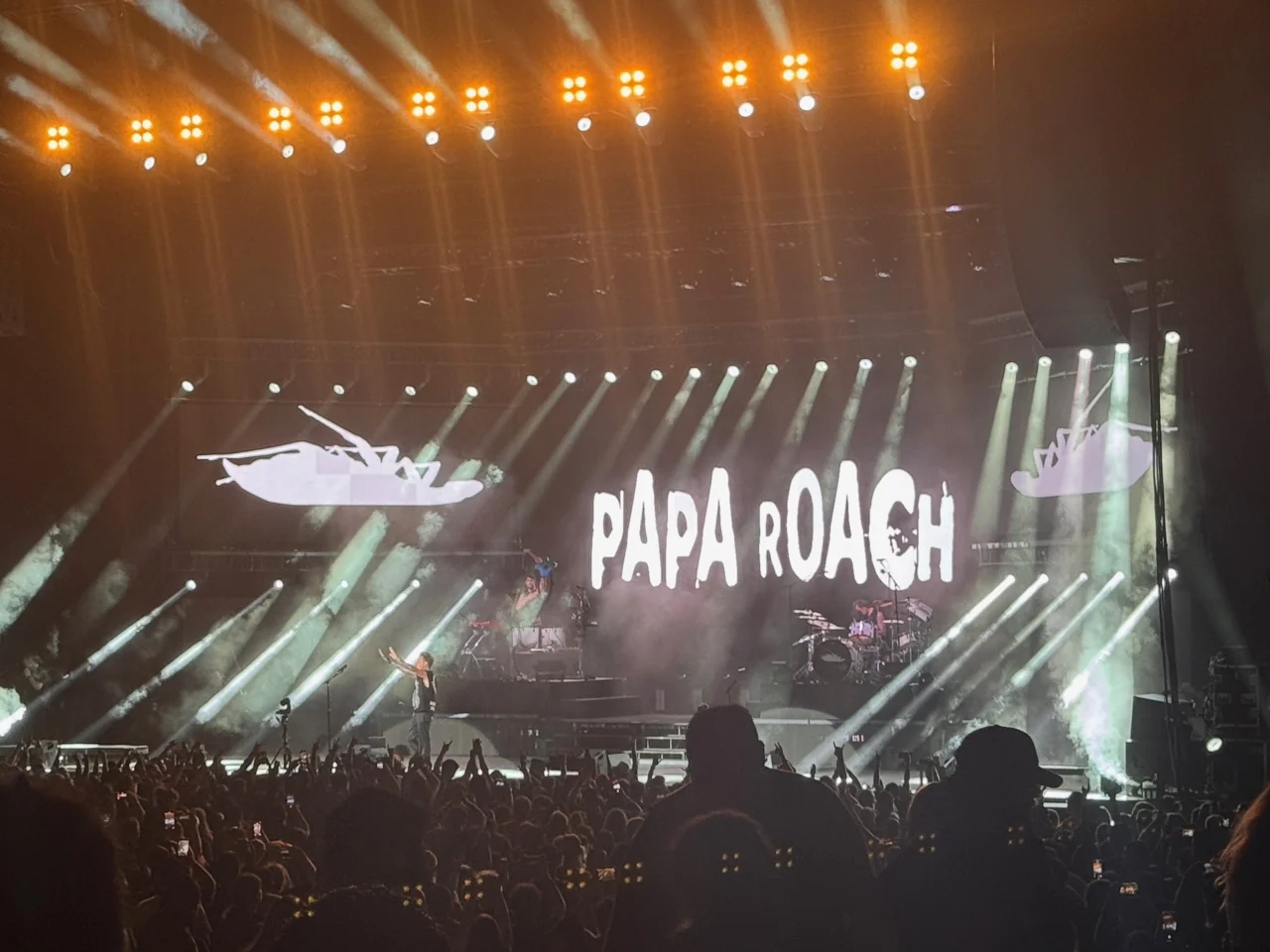 Papa Roach performing beneath white beams and an illuminated band-name backdrop, with raised audience hands in the foreground.
