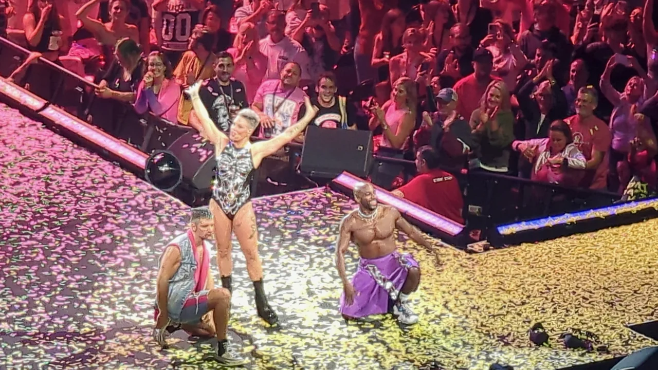 P!nk with arms raised between two dancers on a confetti-covered stage.