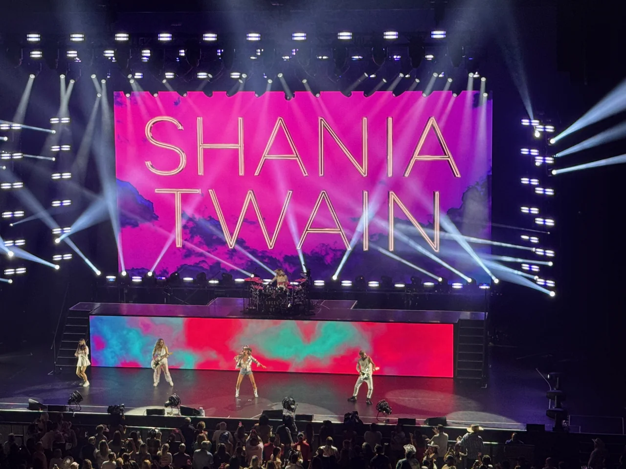 Shania Twain and her band beneath a large pink name backdrop and intersecting white stage beams.