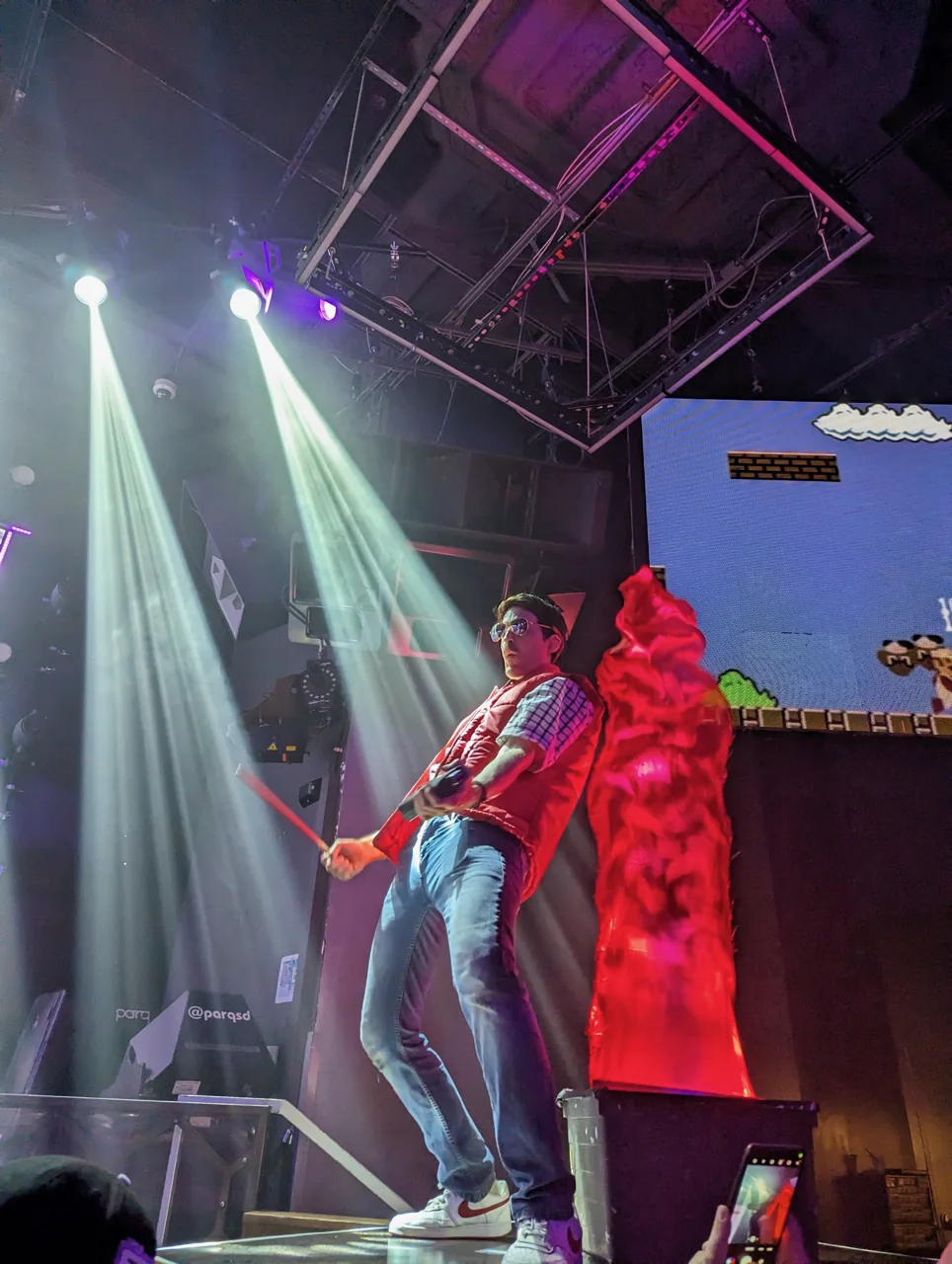A member of The Flux Capacitors in a red vest, checked shirt, jeans, and sunglasses, holding drumsticks beneath white spotlights beside a red fabric flame and a pixel-art game backdrop.