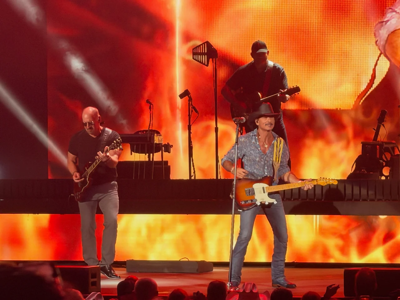 Tim McGraw playing guitar with the band against a fiery orange video backdrop.