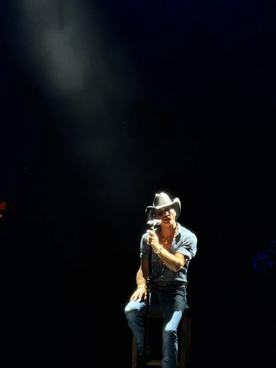 Tim McGraw seated at a microphone under a single spotlight against a dark stage.