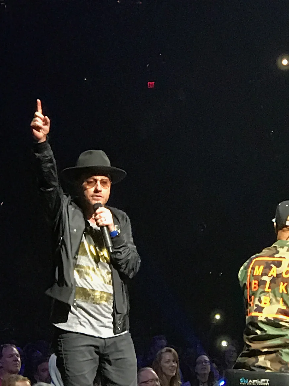 TobyMac wearing a black hat and tinted glasses, holding a microphone and raising one index finger against the dark arena, with audience faces below.