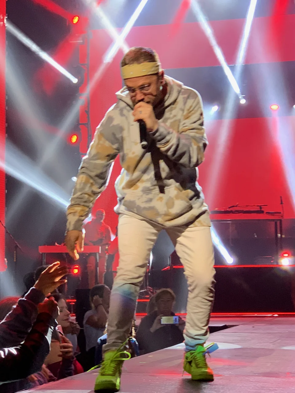 TobyMac leaning forward with a microphone near the audience, wearing camouflage and bright green shoes against red panels and crossing white spotlights.